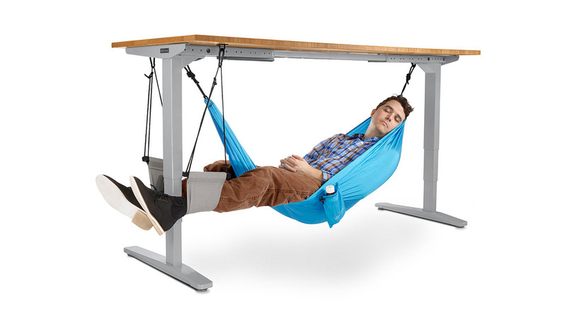 A man snoozes, relaxed in a light blue hammock slung underneath his desk. His eyes are closed and he has a smile on his face. 